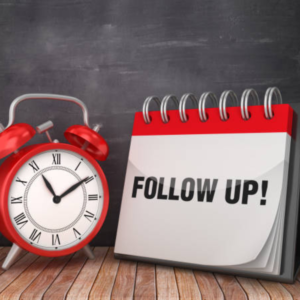 Red alarm clock and desk calendar with "FOLLOW UP!" text on a wooden table against a chalkboard background.